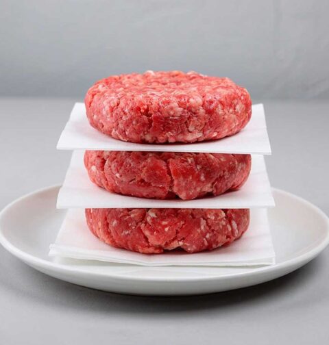 3 nog niet gebakken beefburgers op een wit bordje met vetvrij papier ertussen.
