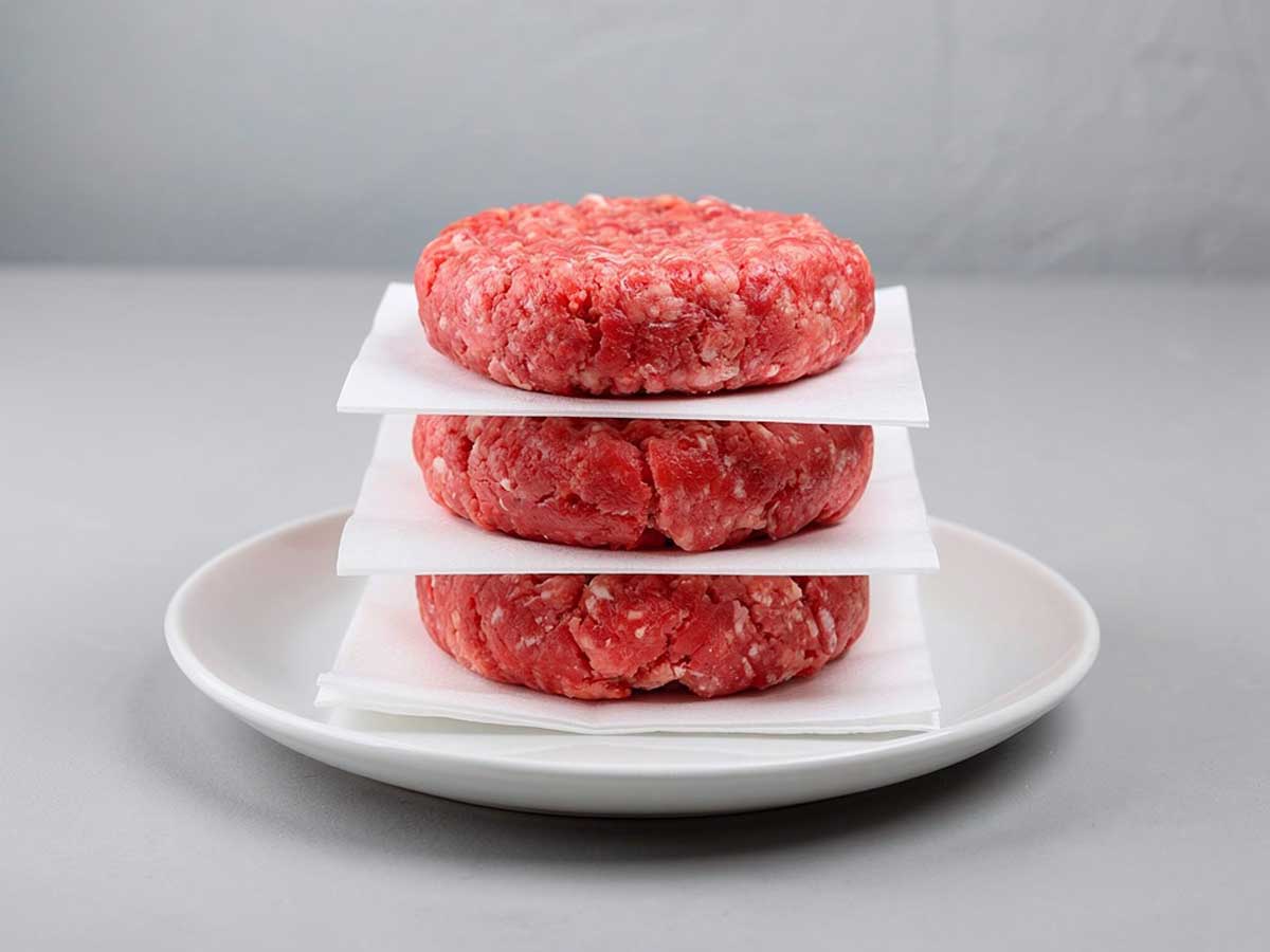 3 nog niet gebakken beefburgers op een wit bordje met vetvrij papier ertussen.