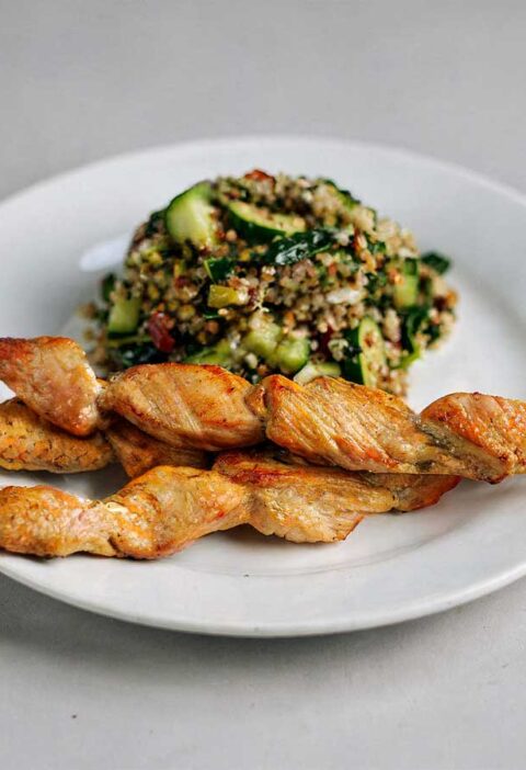 Een wit bordje met groene quinoa-salade en 3 kipspiesjes.