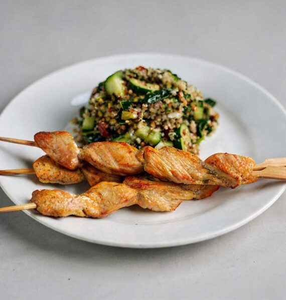 Een wit bordje met groene quinoa-salade en 3 kipspiesjes.