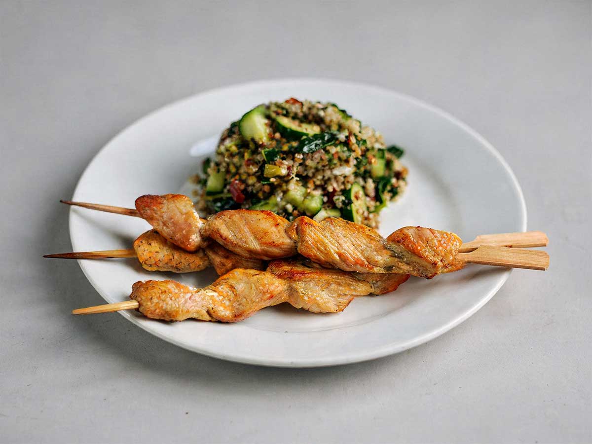 Een wit bordje met groene quinoa-salade en 3 kipspiesjes.