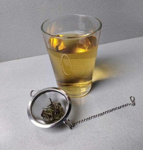 Een glas rozemarijn thee, met daarvoor de resten in een thee ei.
