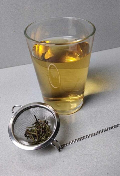 Een glas rozemarijn thee, met daarvoor de resten in een thee ei.
