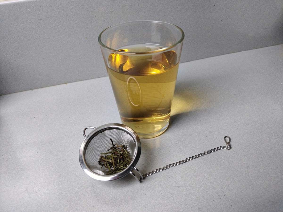 Een glas rozemarijn thee, met daarvoor de resten in een thee ei.