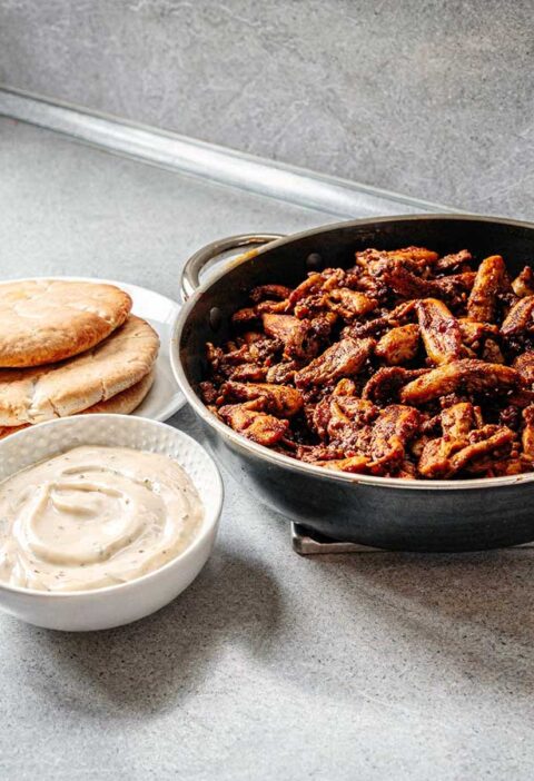 Een koekenpan vol gebakken shoarma, met pitabroodjes en knoflooksaus.