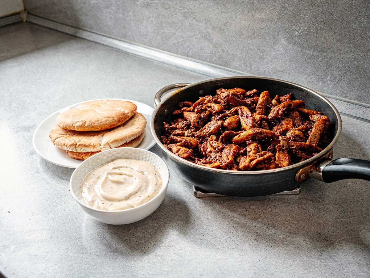 Een koekenpan vol gebakken shoarma, met pitabroodjes en knoflooksaus.