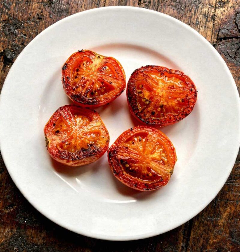 Een wit bord vol met gebakken halve tomaten met de gekaramelliseerde kant boven.