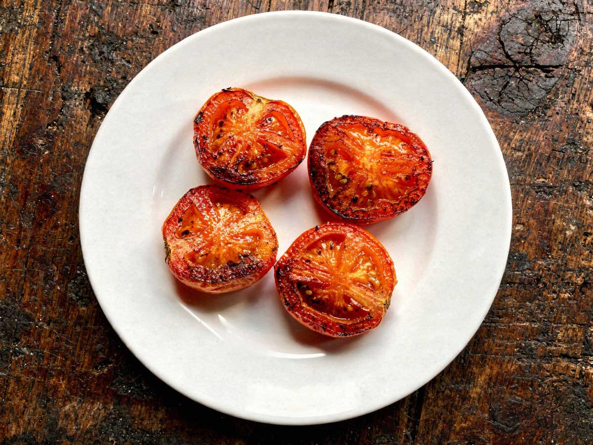 Een wit bord vol met gebakken halve tomaten met de gekaramelliseerde kant boven.