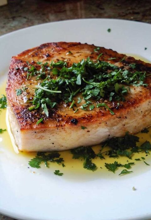 Gebakken zwaardvisfilet op een wit bord met verse groene kruiden en olijfolie