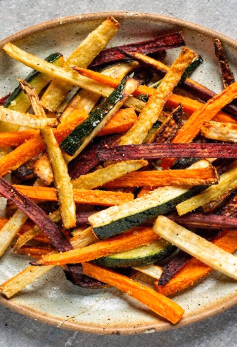 Een grote keramische schaal gevuld met een kleurrijke mix van gebakken groentefrietjes van wortel, biet, courgette en pastinaak.