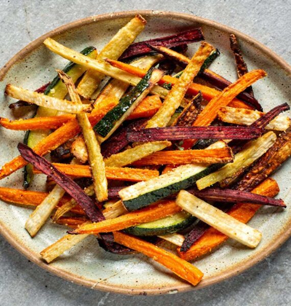 Een grote keramische schaal gevuld met een kleurrijke mix van gebakken groentefrietjes van wortel, biet, courgette en pastinaak.