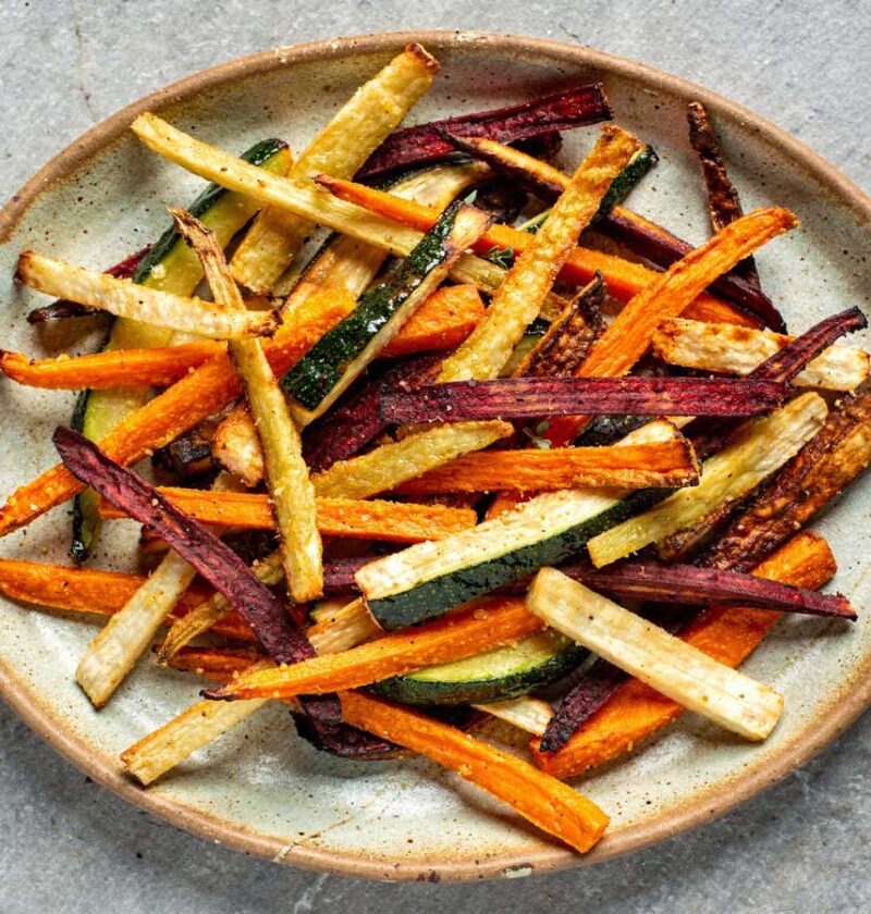 Een grote keramische schaal gevuld met een kleurrijke mix van gebakken groentefrietjes van wortel, biet, courgette en pastinaak.