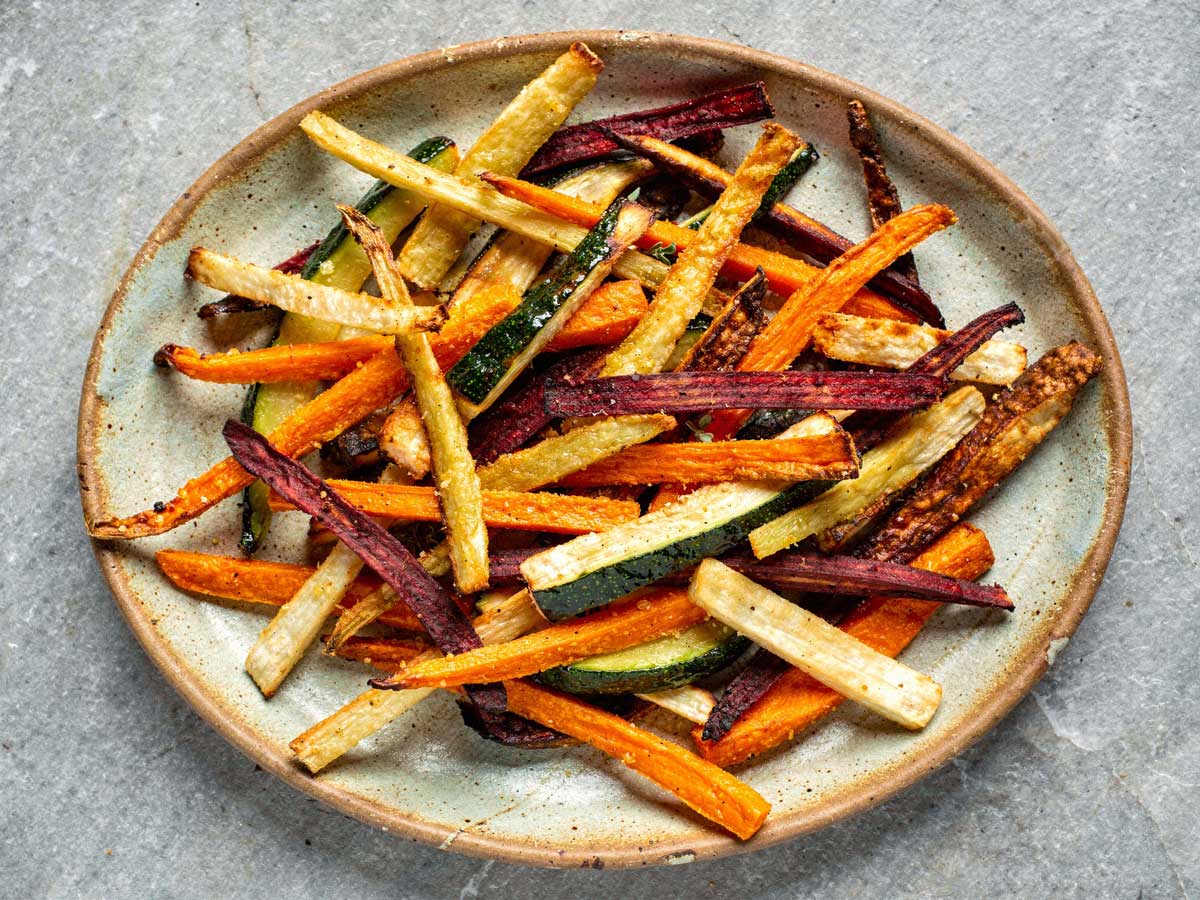 Een grote keramische schaal gevuld met een kleurrijke mix van gebakken groentefrietjes van wortel, biet, courgette en pastinaak.