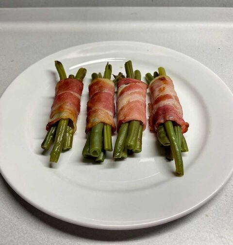 Een wit bord met 4 bundeltjes haricots vert omwikkeld met gebakken spek.