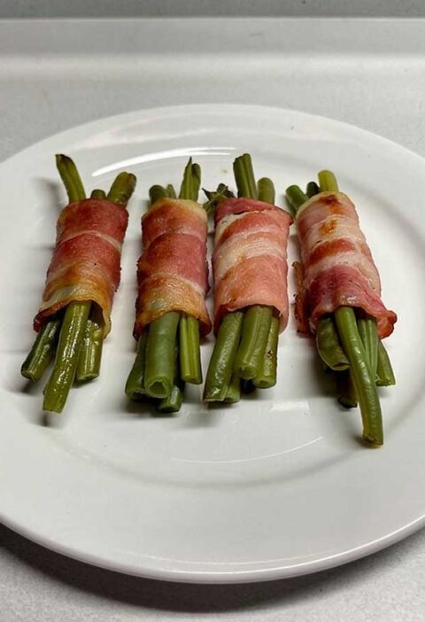 Een wit bord met 4 bundeltjes haricots vert omwikkeld met gebakken spek.