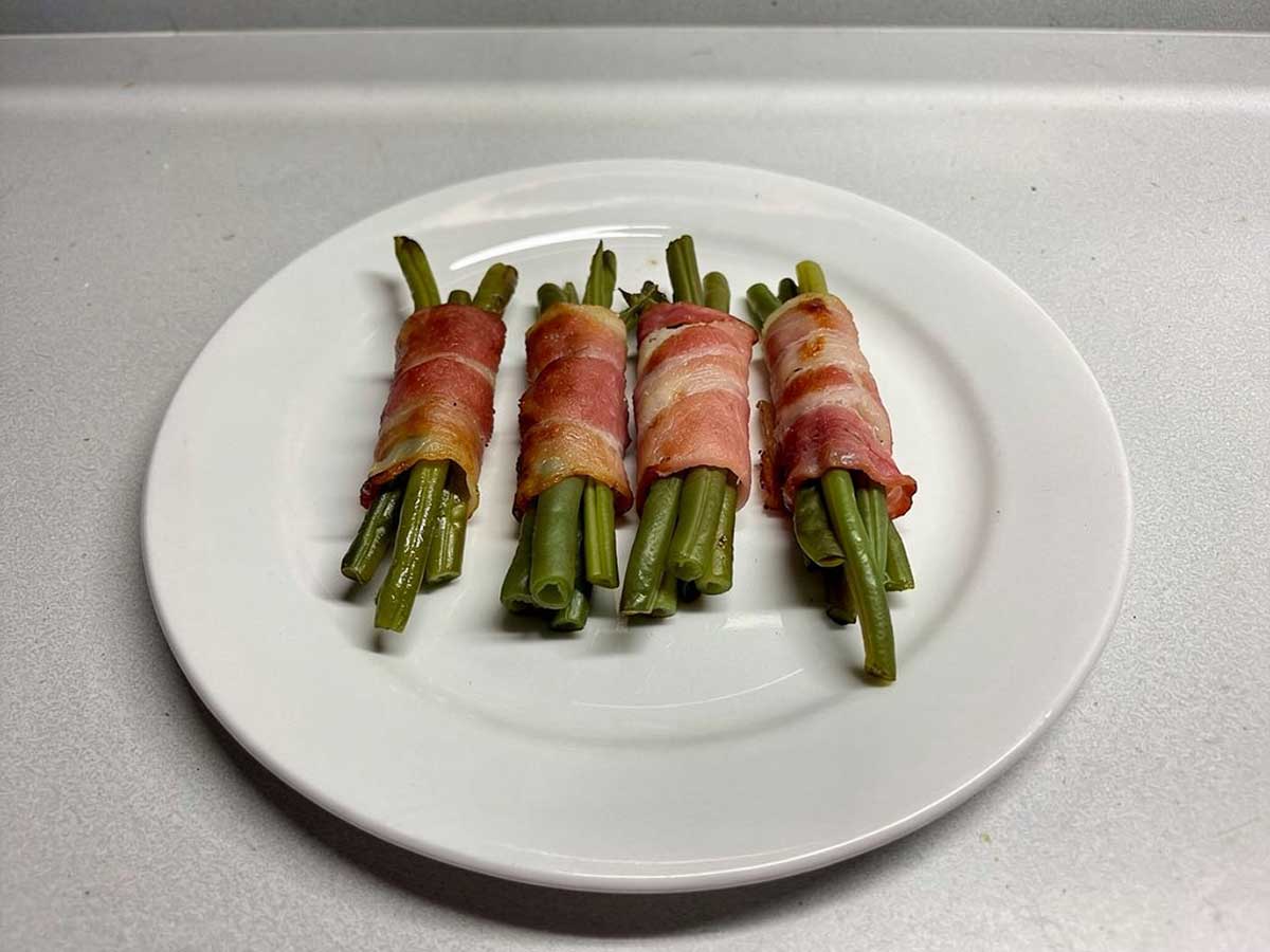 Een wit bord met 4 bundeltjes haricots vert omwikkeld met gebakken spek.