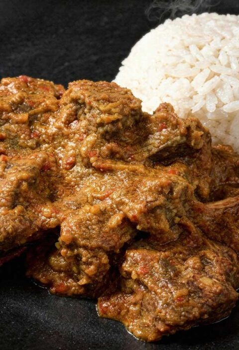 Een portie malse, donkere rundvlees rendang op een plat zwart bord met een bergje witte rijst.