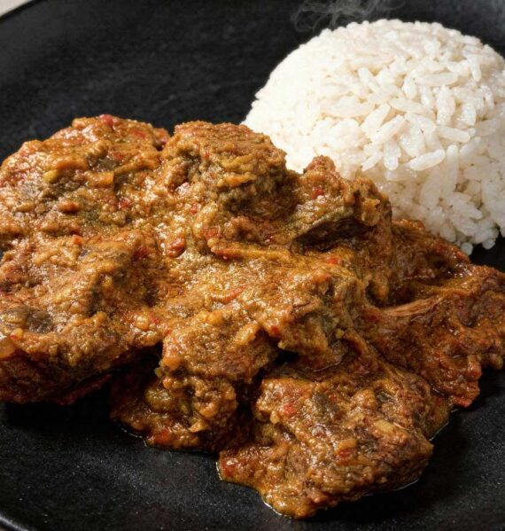 Een portie malse, donkere rundvlees rendang op een plat zwart bord met een bergje witte rijst.