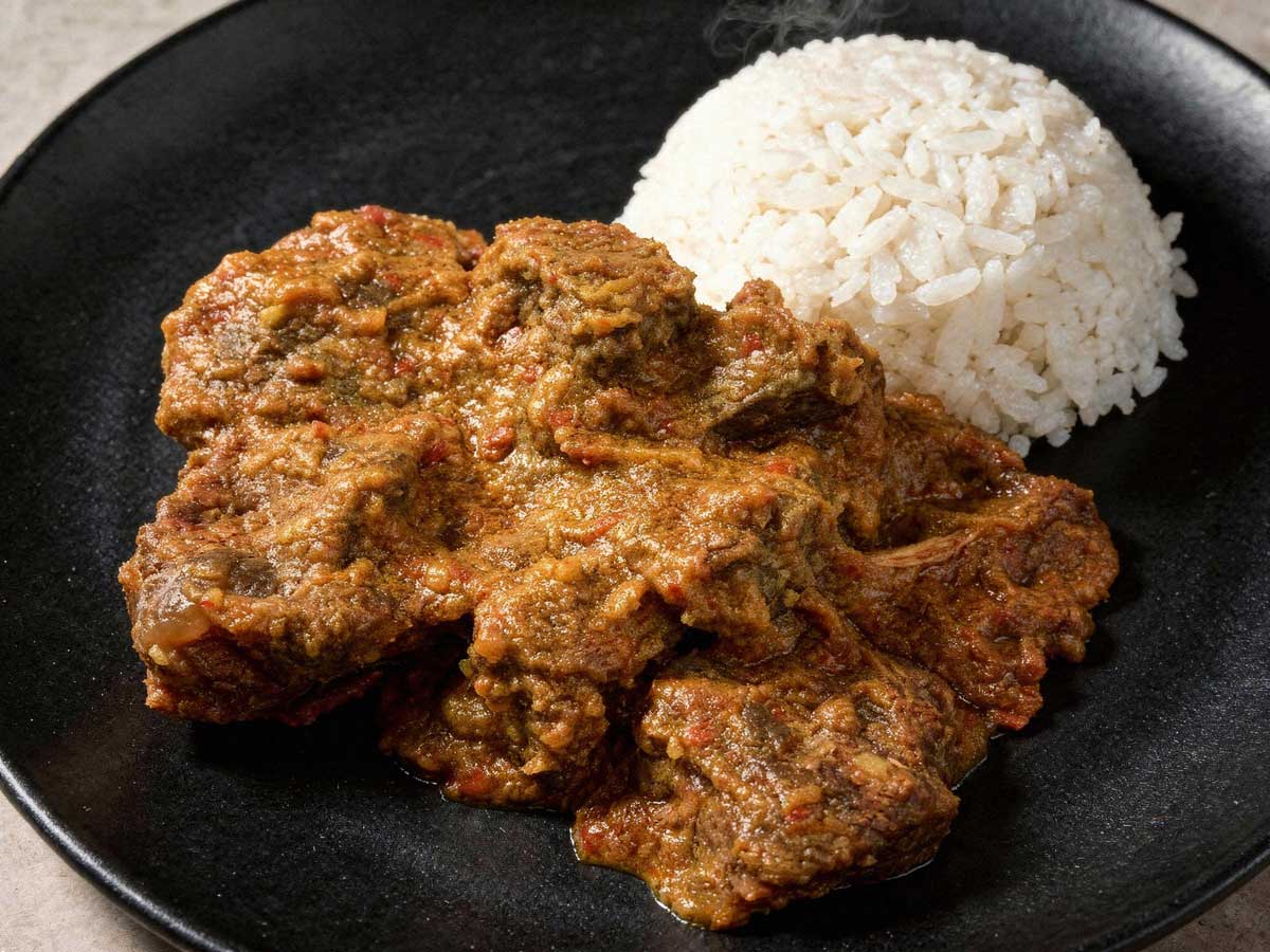 Een portie malse, donkere rundvlees rendang op een plat zwart bord met een bergje witte rijst.