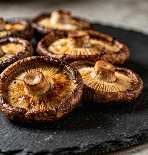 Een stapel sappig gebakken shiitake paddenstoelen met steel, omgekeerd gepresenteerd op een donker leisteen bord.