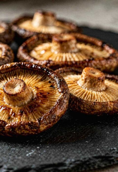 Een stapel sappig gebakken shiitake paddenstoelen met steel, omgekeerd gepresenteerd op een donker leisteen bord.