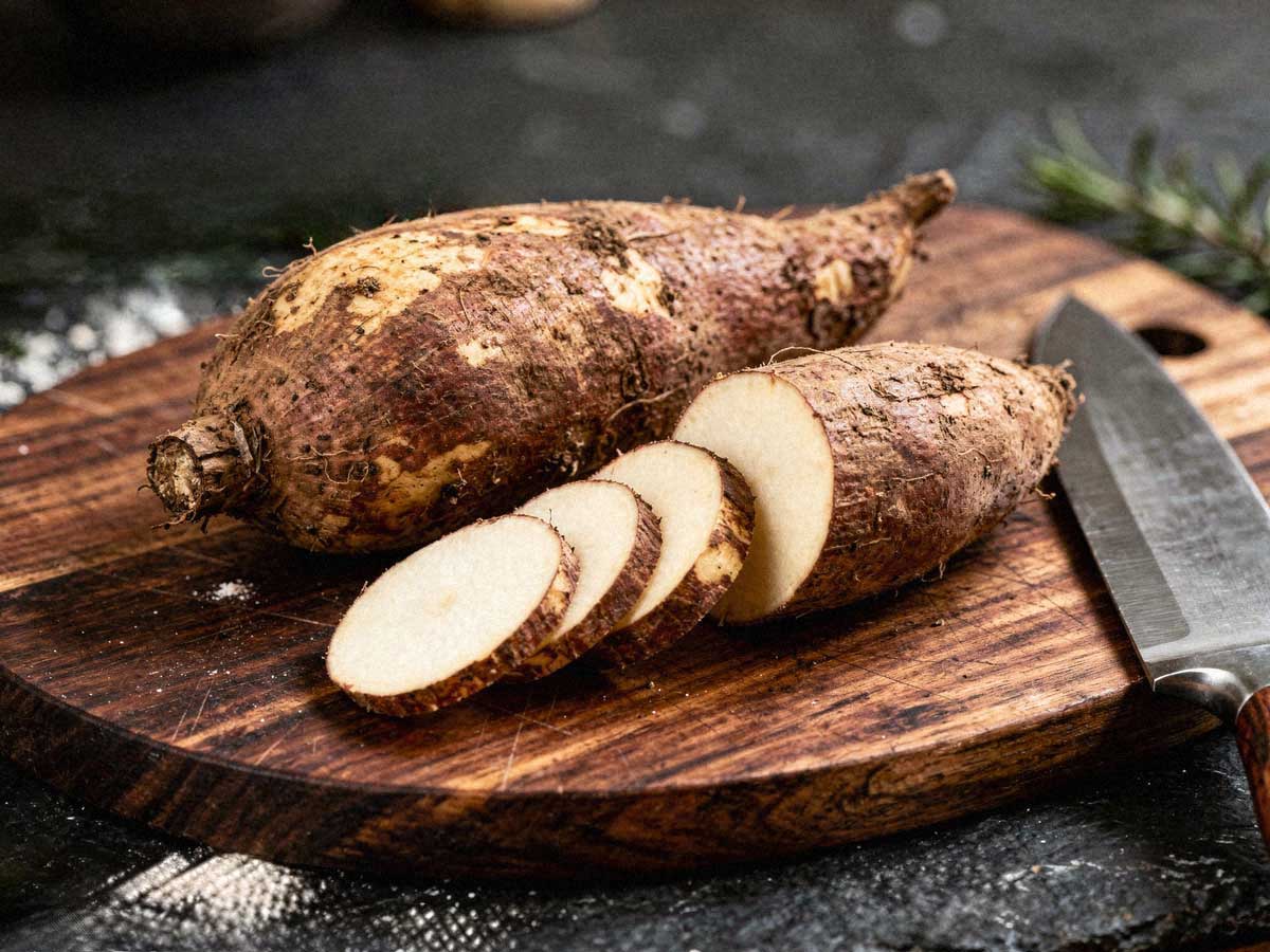 Twee rauwe yams op een snijplank waarvan één deels in plakken is gesneden.