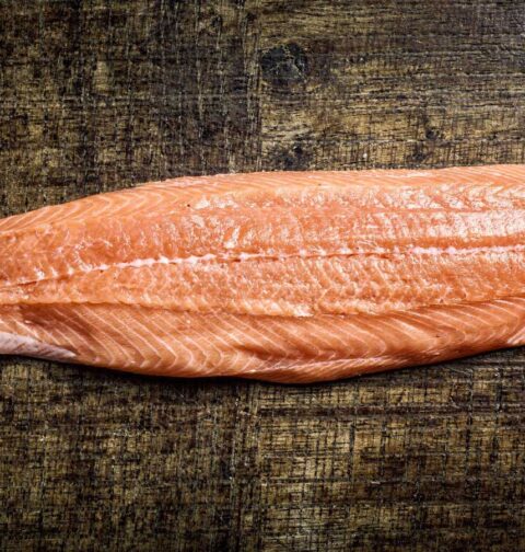 Een dik stuk rauwe roze zalmfilet zonder huid met witte vetlijnen op een donkere houten tafel. Hiervan ga ik zalmrollade maken.