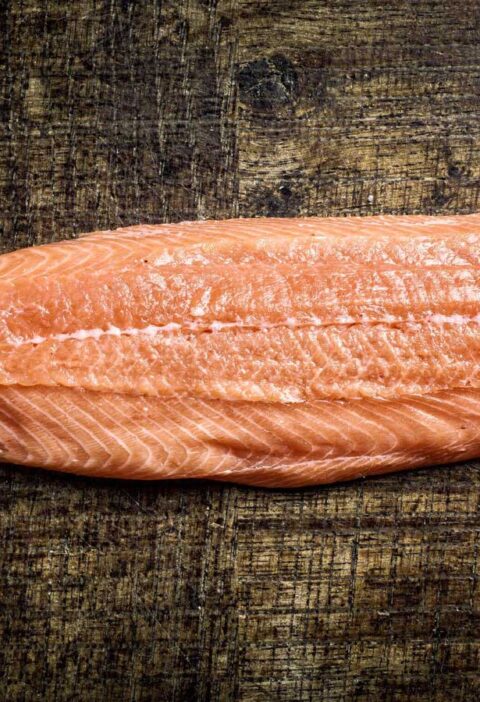 Een dik stuk rauwe roze zalmfilet zonder huid met witte vetlijnen op een donkere houten tafel. Hiervan ga ik zalmrollade maken.