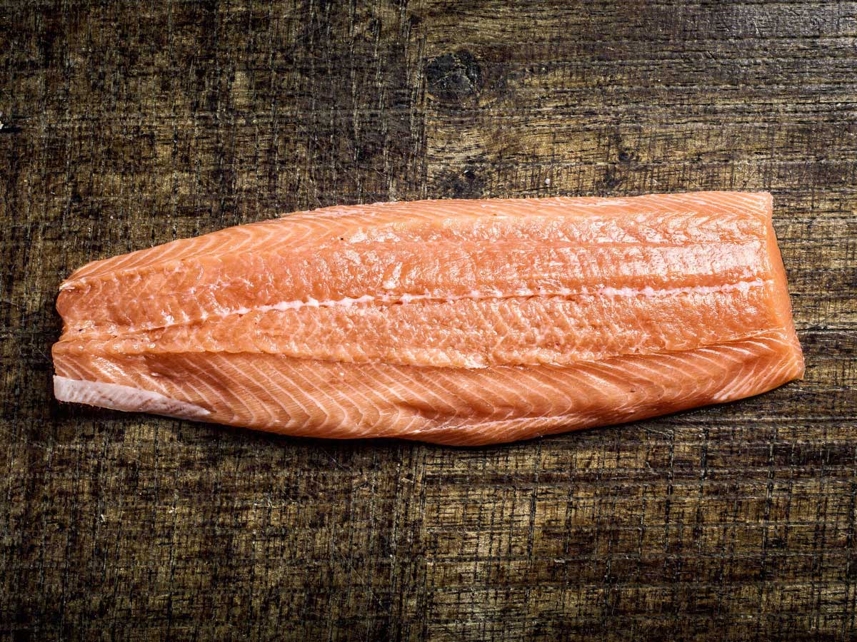 Een dik stuk rauwe roze zalmfilet zonder huid met witte vetlijnen op een donkere houten tafel. Hiervan ga ik zalmrollade maken.