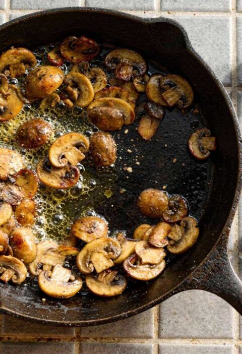 Gebakken plakjes kastanjechampignons bruisend in de boter in een gietijzeren pan met rauwe champignons ernaast