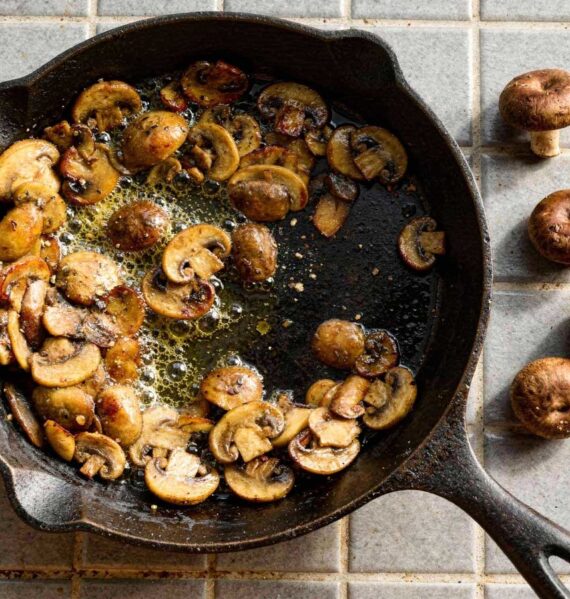 Gebakken plakjes kastanjechampignons bruisend in de boter in een gietijzeren pan met rauwe champignons ernaast