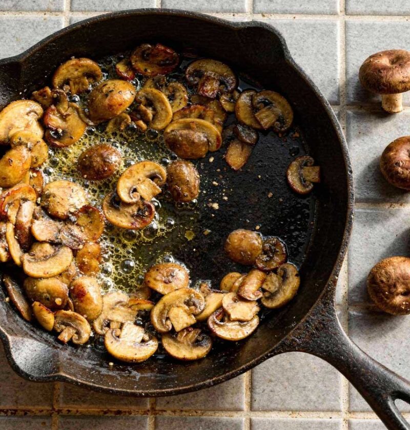 Gebakken plakjes kastanjechampignons bruisend in de boter in een gietijzeren pan met rauwe champignons ernaast