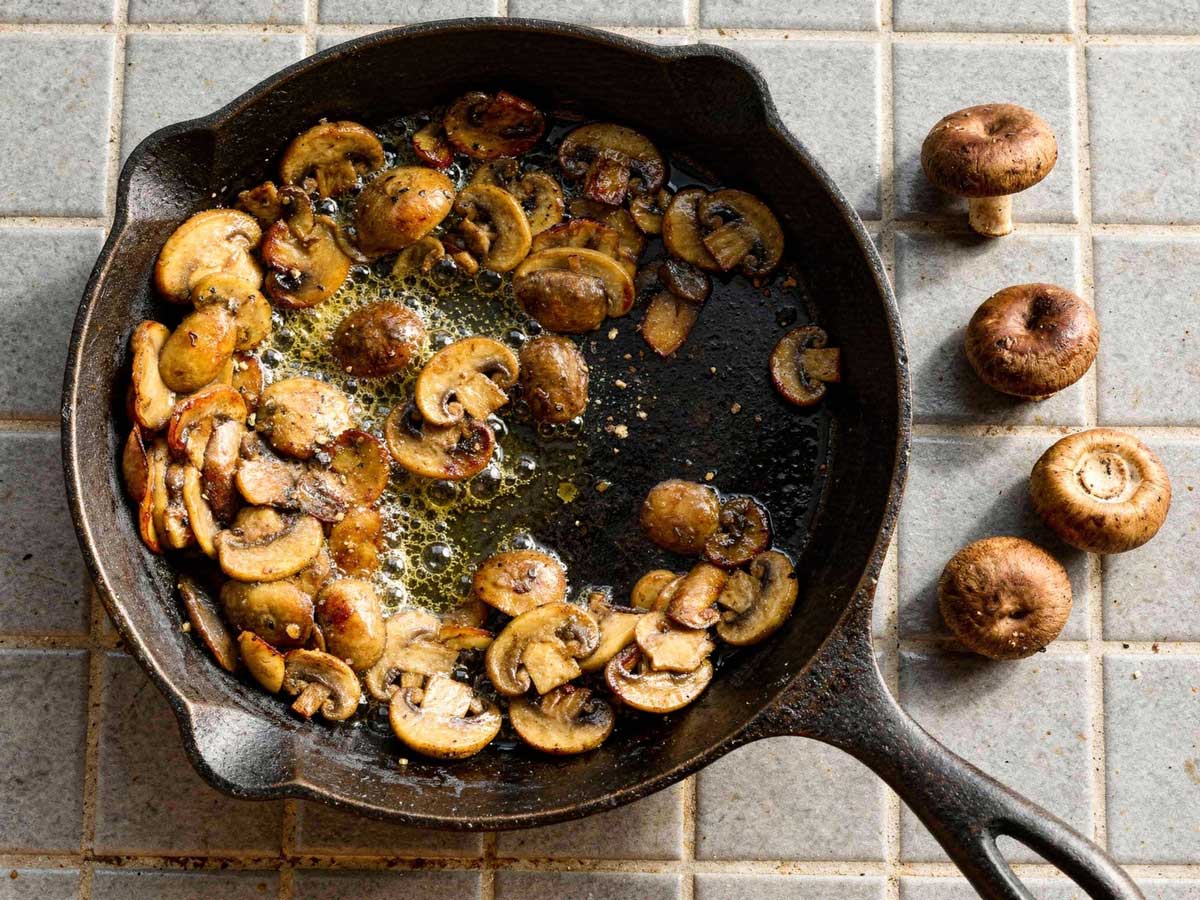Gebakken plakjes kastanjechampignons bruisend in de boter in een gietijzeren pan met rauwe champignons ernaast