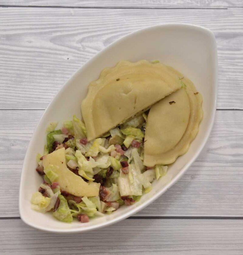 Een heerlijke groene salade met appel, spek en verse ravioli, in een wit schaaltje.
