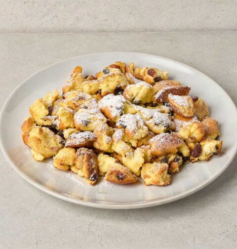 Een wit bord met luchtige Kaiserschmarrn, royaal bestrooid met poedersuiker.