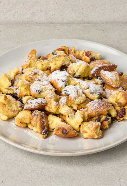 Een wit bord met luchtige Kaiserschmarrn, royaal bestrooid met poedersuiker.