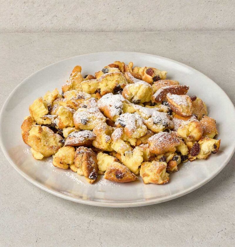 Een wit bord met luchtige Kaiserschmarrn, royaal bestrooid met poedersuiker.