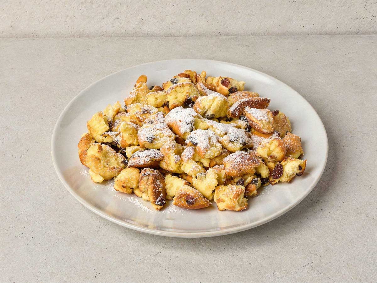 Een wit bord met luchtige Kaiserschmarrn, royaal bestrooid met poedersuiker.