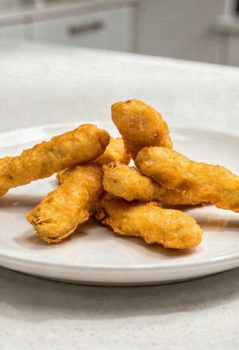 Vijf goudbruin gefrituurde kipfingers netjes geserveerd op een wit bord.