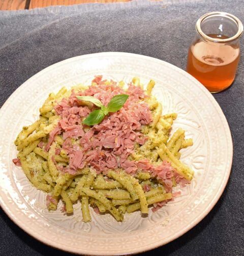 Een lichtgrijs bord met daarop een heerlijke macheroni met pistachepesto en licht krokante mortadella.