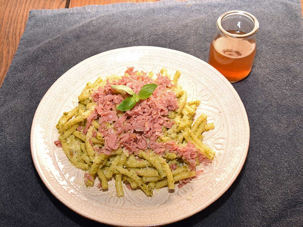Een lichtgrijs bord met daarop een heerlijke macheroni met pistachepesto en licht krokante mortadella.
