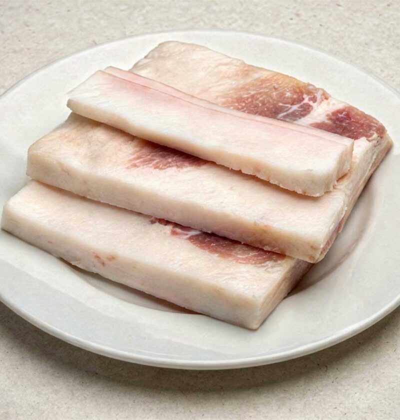 Twee grotere stukken gebakken vet spek met twee kleinere plakken ongebakken spek op een witte platte schaal.