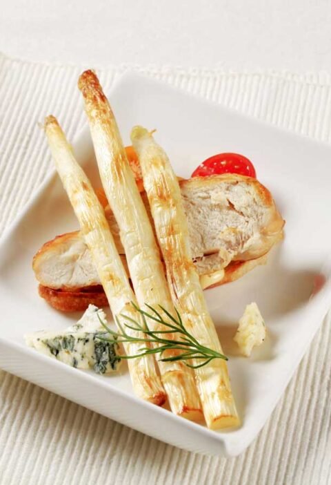 3 gebakken witte asperges op een bord met onder andere een stuk gebakken kip.